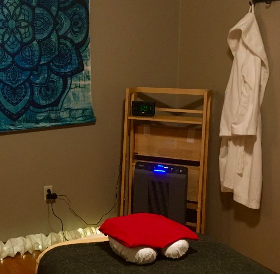 Color image of massage table with beige linens and a charcoal blanket and a white face cradle on top with a red cover. Back corner is an air purifier and clock in a light wood bookcase. Right wall has a clean, available terrycloth spa robe. Left is a predominately blue weaving.