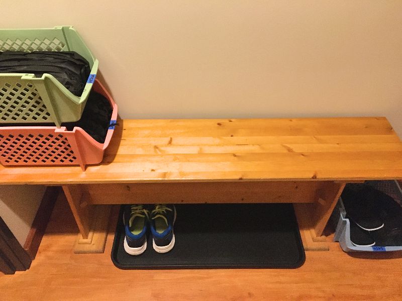 Color photo of a shoe bench with a green and pink basket on the left with bagged, clean spa slippers, a black shoe mat under the wooden bench with blue tennis shoes on it, and a blue basket on the right for used slippers